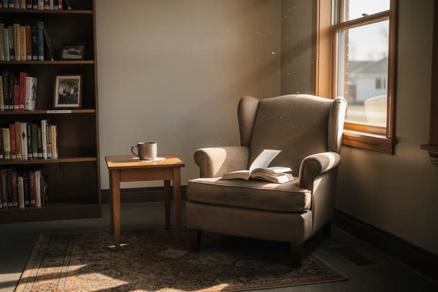 A genuinely quiet empty neighborhood library reading nook in mid-afternoon. No human figures. One comfortable worn armch