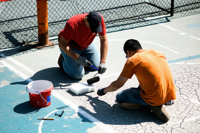 A tender practical repair on a sun-baked community basketball court during bright late morning. Two adults kneel on the