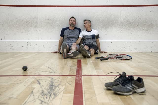 A genuinely quiet empty squash court after casual evening play. Two adult friends sit side by side against the back wall