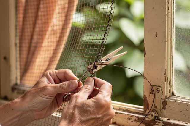 An intimate domestic repair beside an open wooden sash window. One resident uses a curved needle and dark waxed thread t