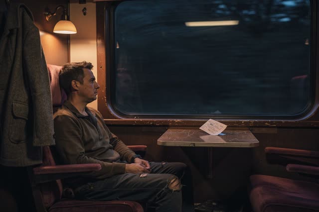 A genuinely quiet overnight train compartment. One adult passenger sits comfortably beside a dark window, awake but doin