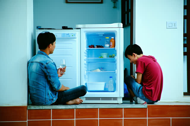 A genuinely quiet small apartment kitchen during a humid night. Two adult friends sit on the cool glazed-tile floor besi
