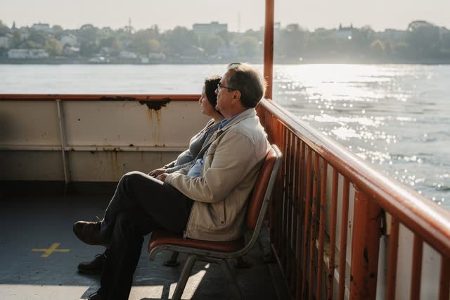 I dream of the open side deck of a modest commuter ferry in low clear morning light. Two adult passengers sit comfortabl