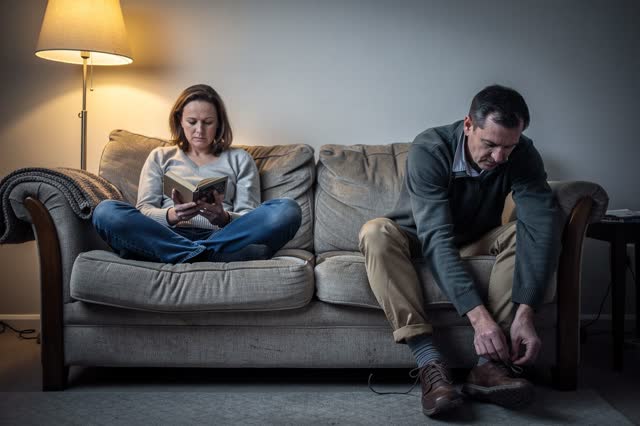 I dream of two adults resting in a modest living room after an ordinary long day. They sit in familiar unequal indentati