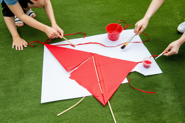 I dream of two adults repairing a torn red paper kite at the edge of a grassy park beneath a plain picnic shelter. Use a