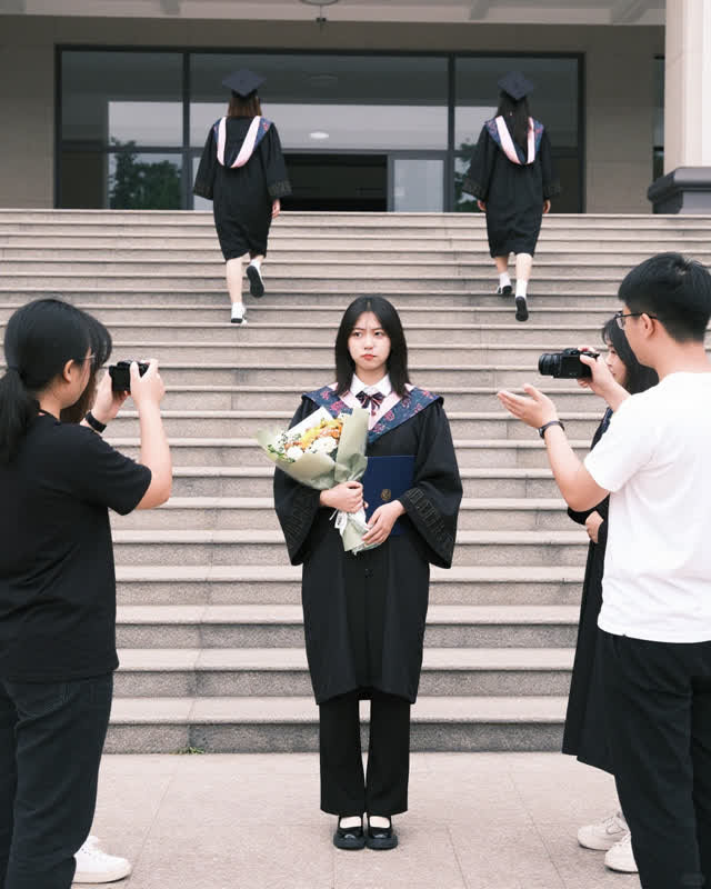 A low-tension social challenge on broad pale school steps after graduation. One adult graduate stands near the center ho
