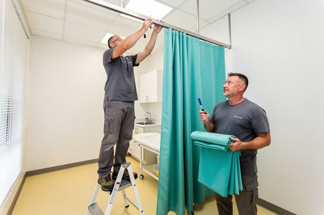 I dream of a bright community clinic exam room where two adult maintenance workers repair a jammed privacy curtain befor