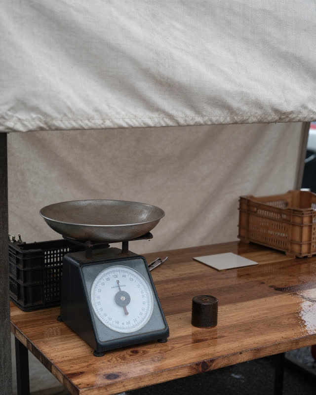 I dream of a small produce stall after rain where the tare weight is still on the pan. Under a pale canvas awning, a pla