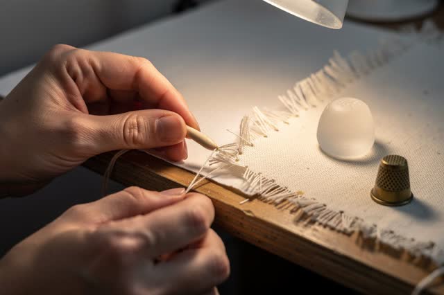I dream of two natural hands at a small mending bench, stitching a torn cream paper edge to a thin cloth backing with pa