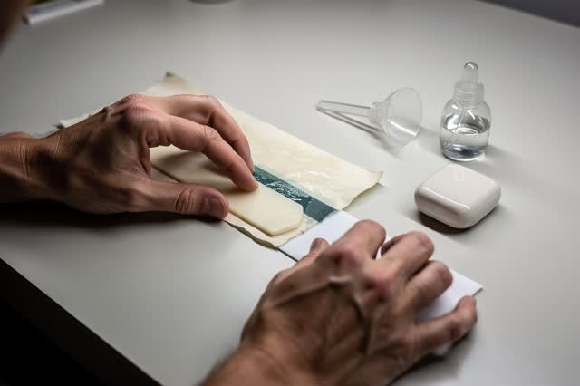 I dream of two natural hands at a clean margin bench, giving a folded pasted tab a softer edge before the blue-green pig