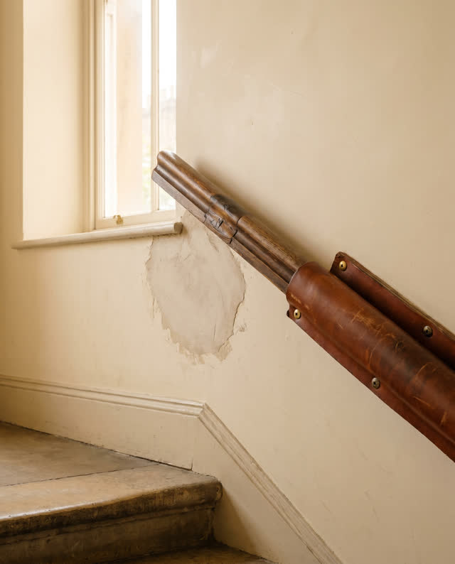 I dream that the landing teaches the hand where to go. On a small public stair landing, a repaired wooden rail, a feathe
